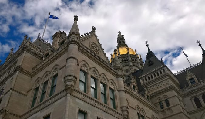 Ornate government building with a blue flag and a gold dome, against a cloudy blue sky