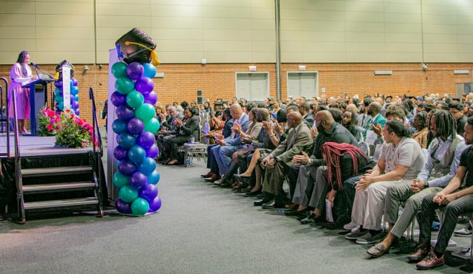 People seated in front of a stage with a person speaking and balloons.