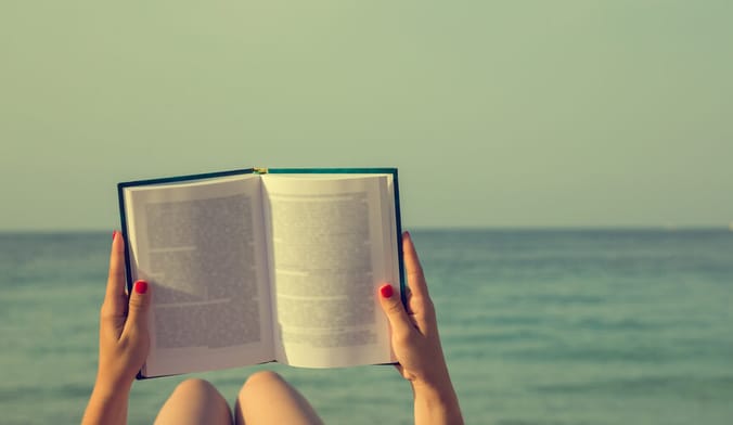 A person's legs and arms are shown reading a book next to a body of water