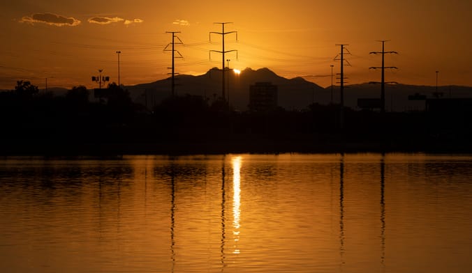 The sun rises behind a mountain range. Powerlines are shown in silhouette. A body of water is in the foreground.