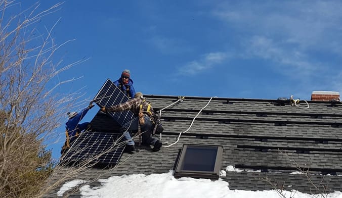 Three people hoist a solar panel onto a roof against a blue sky