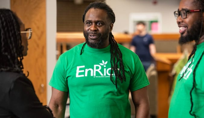 A person talks with two other people. He wears a green t-shirt with the words "EnRich"