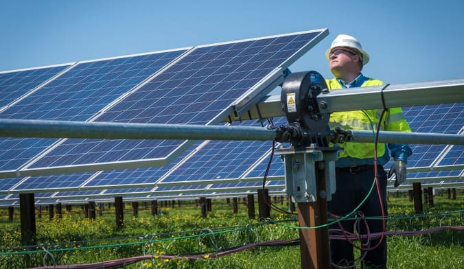 A person in a hard hat and neon yellow vest stands next to a row of solar panels outside in a field
