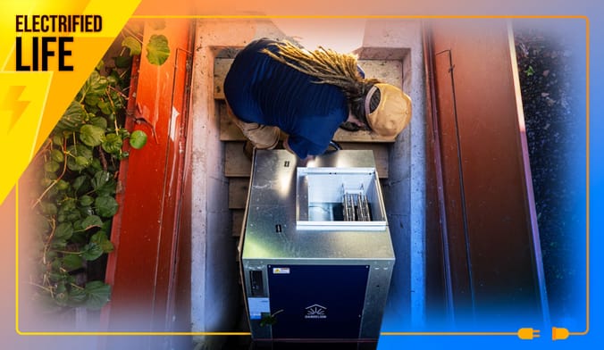 Worker with dreadlocks and yellow cap helps carry geothermal heat pump unit down stairs behind double doors.