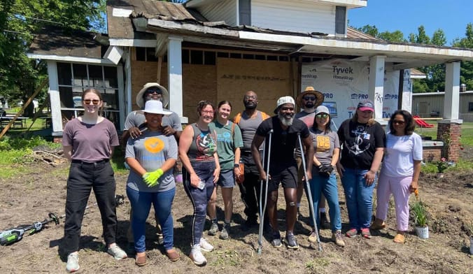 A group of people stand on a patch of dirt in front of a boarded up home