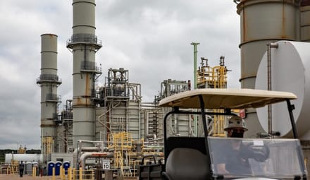 Industrial facility pipes rise up against a cloudy sky. A person sits in a golf cart in the foreground.