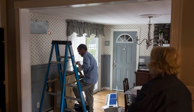 A person stands next to a ladder inside a home. An appliance lays on the ground nearby.