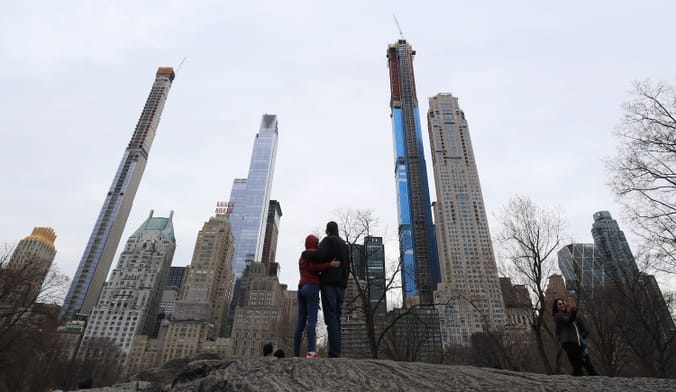 Two people with their arms around each other stand in a park looking at tall city buildings.