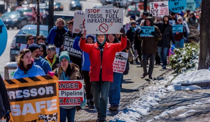 People in winter clothing line up on a sidewalk and hold anti-fossil-fuel signs