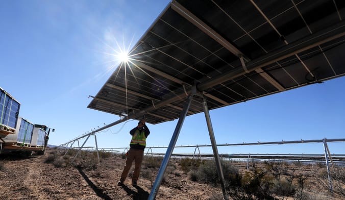 A person in a yellow vest works on ground-mounted solar panels viewed from below