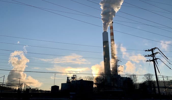Smoke billows out of pipes and into the sky. Power lines are silhouetted against the sky.
