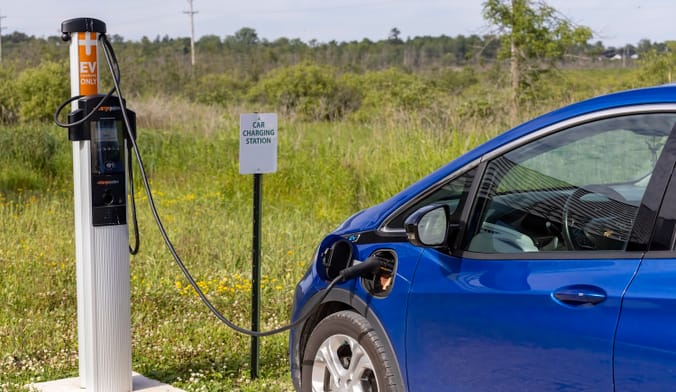 A blue car plugged into an EV charger in front of a large grassy field