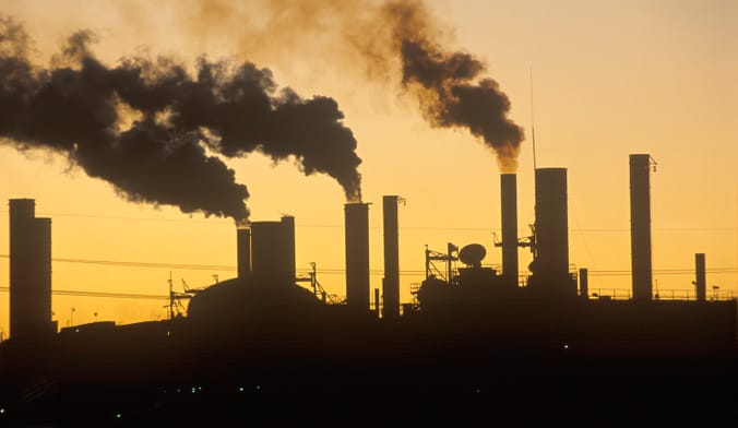 Smoke billows out of industrial buildings silhouetted against the sky at sunset.