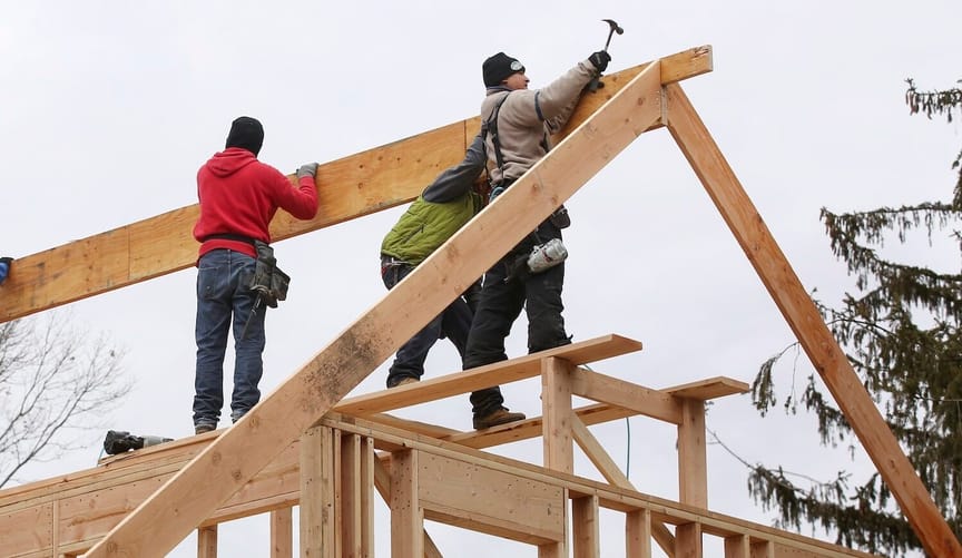 Three people stand on wooden framing for a house. One holds a hammer
