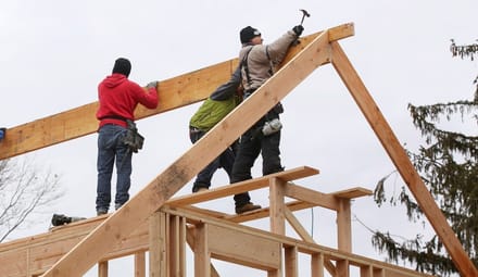 Three people stand on wooden framing for a house. One holds a hammer