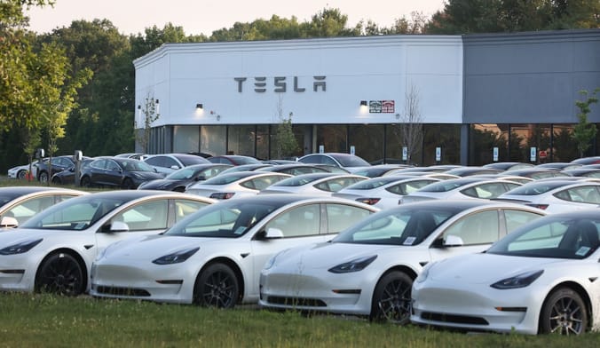 Rows of cars lined up in front of a blocky building with the word "Tesla" written on the gront
