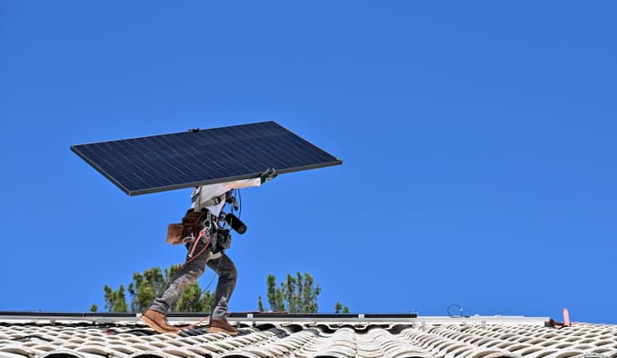 A person on a roof carries a large solar panel on their shoulders