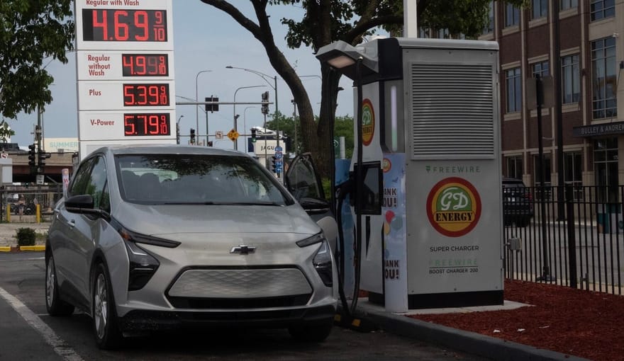 A car parked next to a boxy piece of charging equipment. Behind the car is a large sign displaying gas prices.