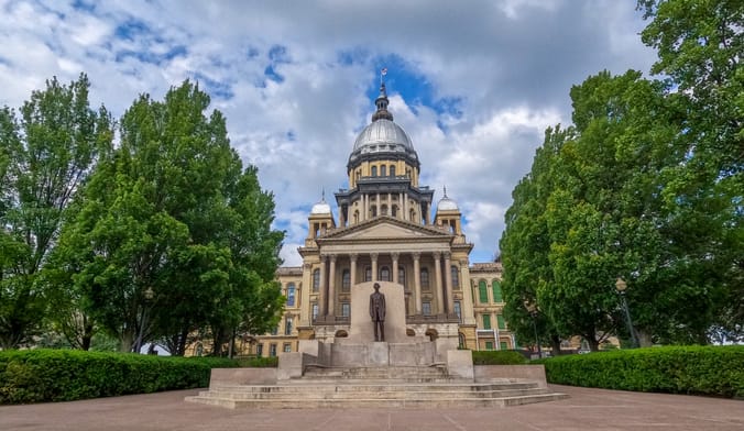 Front view of Illinois State Capitol Building with statue of Abraham Lincoln stands in front. Cloudy blue skies overhead.