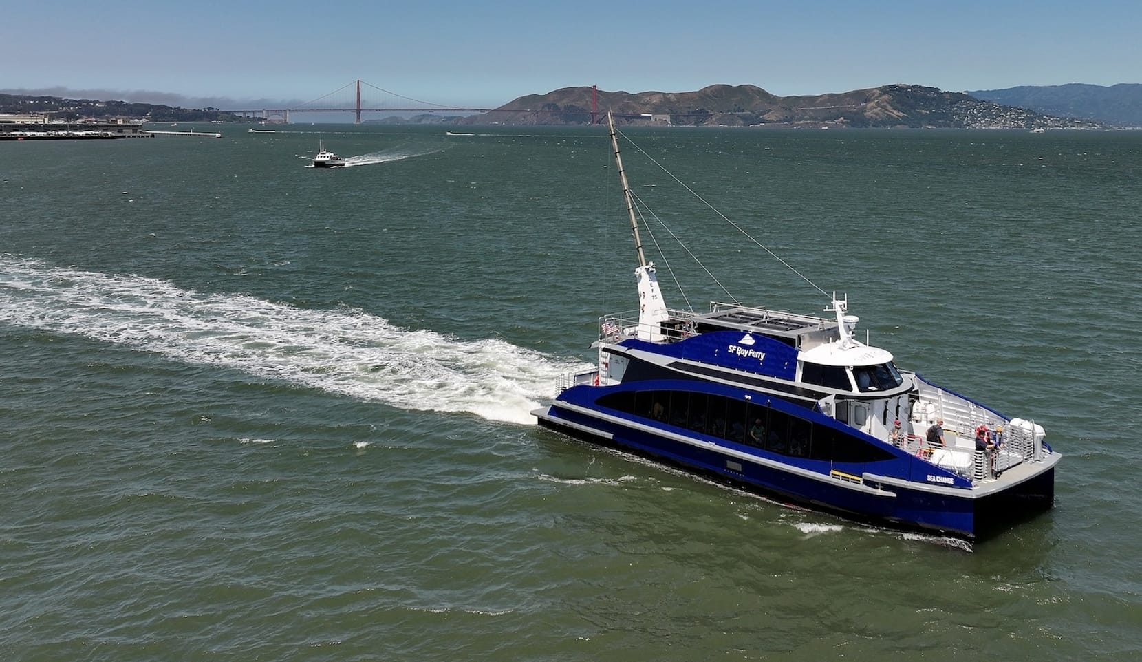 A boat that says "SF Bay Ferry" drives through the water in front of hills and a large bridge.