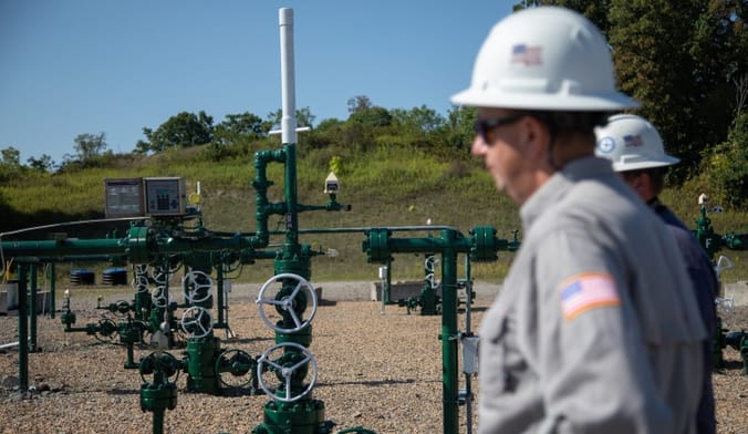 A person in a shirt and hard hat stand in front of piping equipment that feeds underground