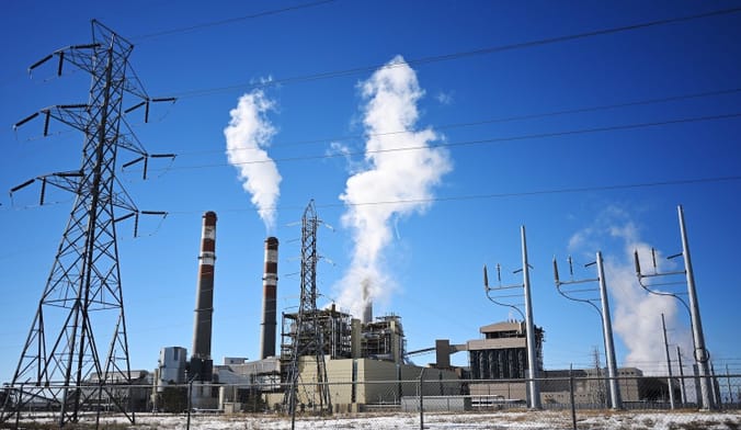 Smoke spews out of an industrial facility against a blue sky. Next to the plant is a transmission tower.