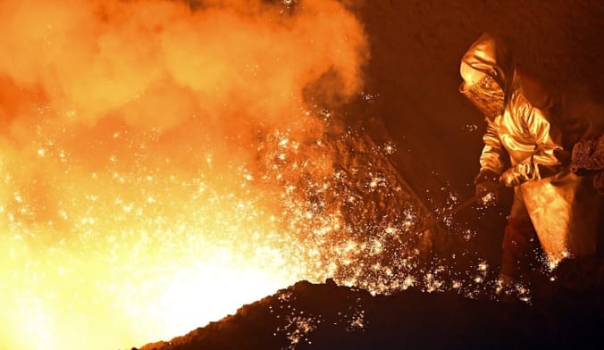 A worker in protective gear stands next to a fiery area with sparks