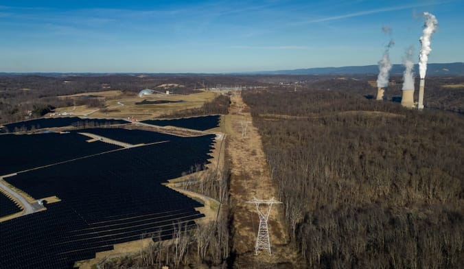 Aerial shot of a transmission line running alongside rows of solar panels and an industrial facility spewing smoke/steam