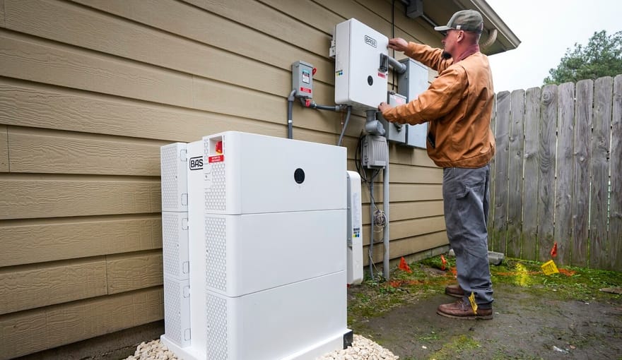 A person in casual clothing stands next to electrical equipment outside of a house