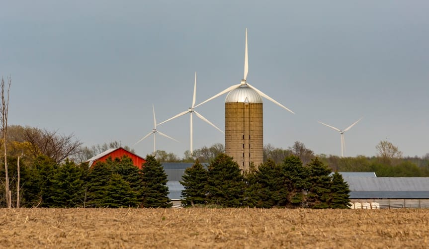 Turbines rise against the sky behind a field with a barn and grain silo