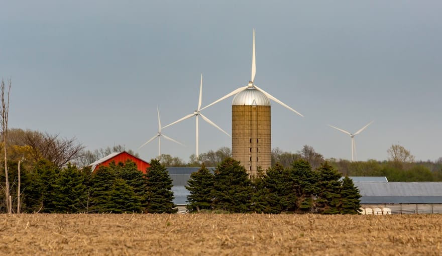 Turbines rise against the sky behind a field with a barn and grain silo