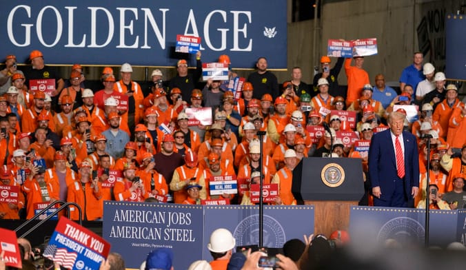 A person in a suit stands on a stage in a sea of people wearing orange vests and hard hats. A large sign reads "Golden Age"