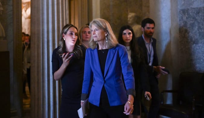 A woman in a blue blazer walks through a government building. She is flanked by four people in business attire.