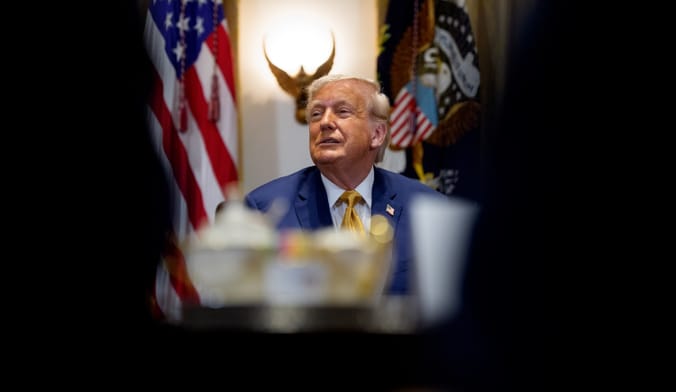 A person in a suit sits at a desk in front of a U.S. flag