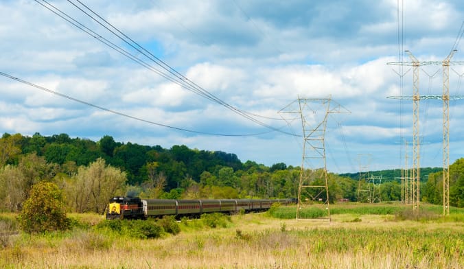 A train runs through a rural field alongside large transmission towers and power lines