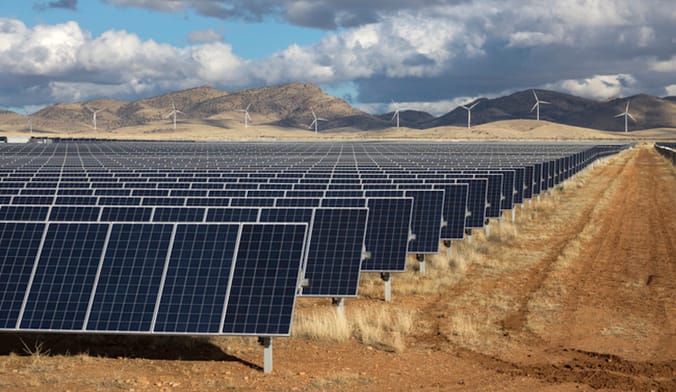 Solar panels sit are installed in a large field. In the background are hills and wind turbines against a cloudy sky.