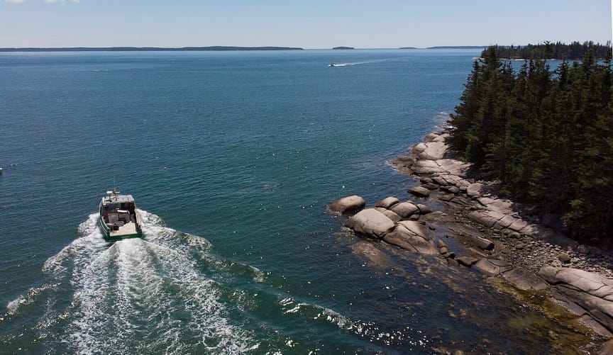 Aerial shot of boat driving through ocean water next to a rocky coast lined with trees
