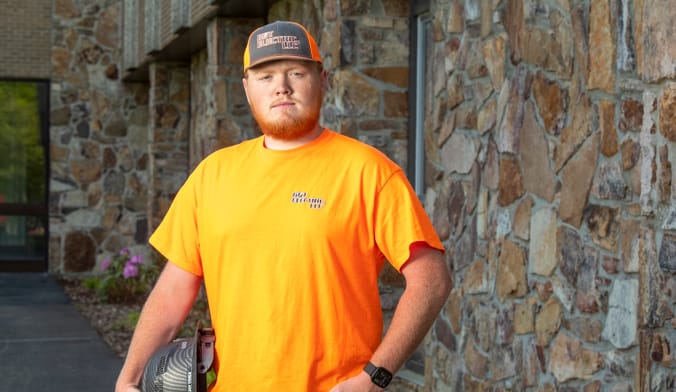 A person in an orange tee shirt holds a hard hat in front of a stone wall.