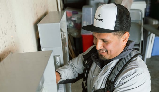 A smiling person in a hat that reads "Haven" looks closely at equipment on a wall