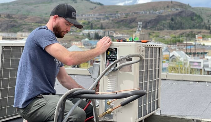 A person in tee shirt and baseball cap works on a heat pump unit on a roof with hills and other buildings in the background