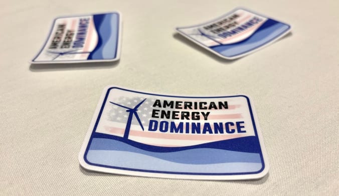 Three stickers on a table. Each has a wind turbine, a U.S. flag, and the words "American Energy Dominance"