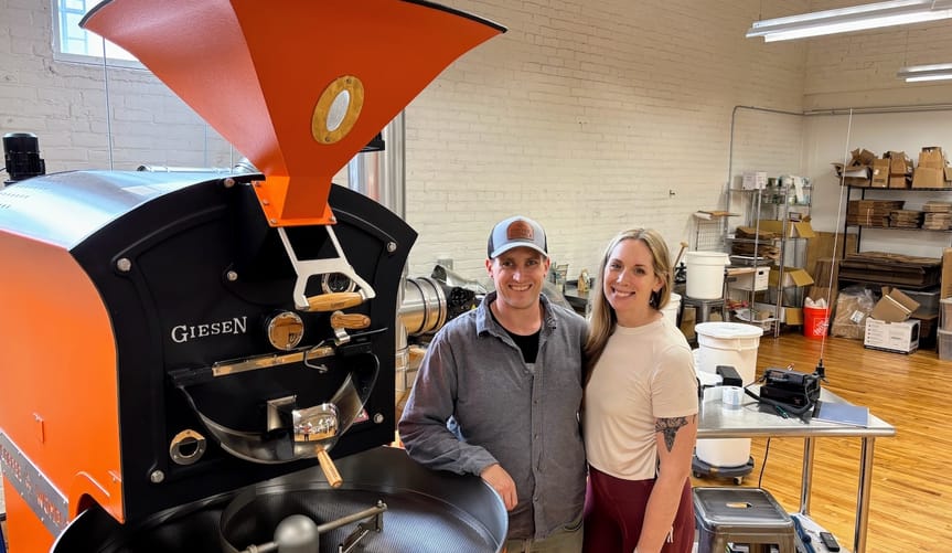 Two people stand next to a large piece of equipment with a funnel affixed to the top.