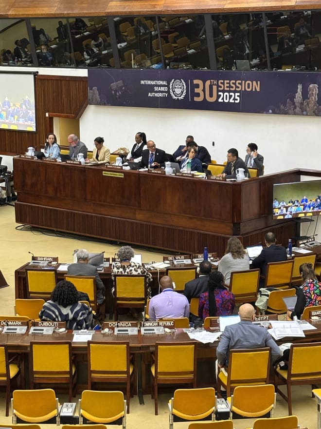 People in business attire sit at long tables facing a panel of people sitting at a raised wooden desk