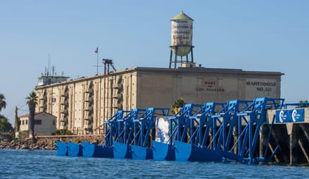 Blue steel structures sit in the water at a wharf with a warehouse on it
