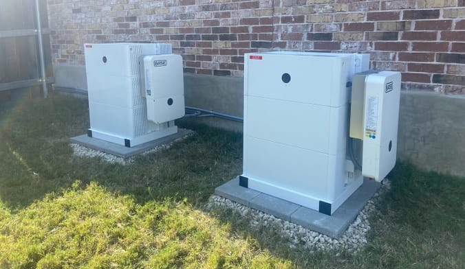 Two large boxy pieces of battery equipment sit outside next to a brick wall