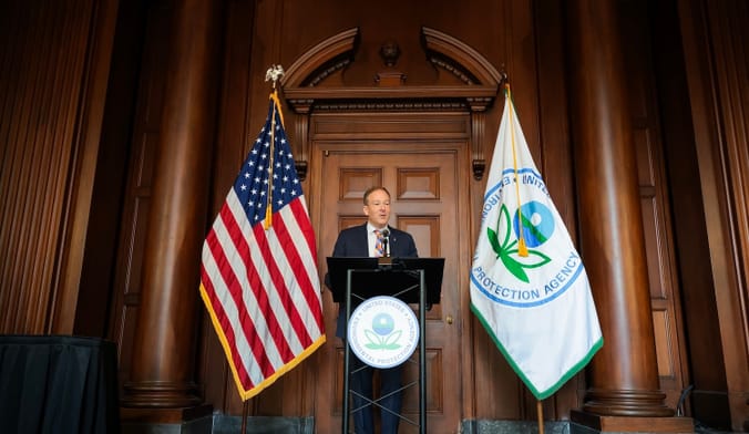 A person speaks into a microphone at a podium, flanked by the U.S. flag and the Environmental Protection Agency flag.