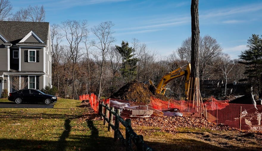 A fence runs between a house with a car parked in front and a patch of land with construction equipment on it