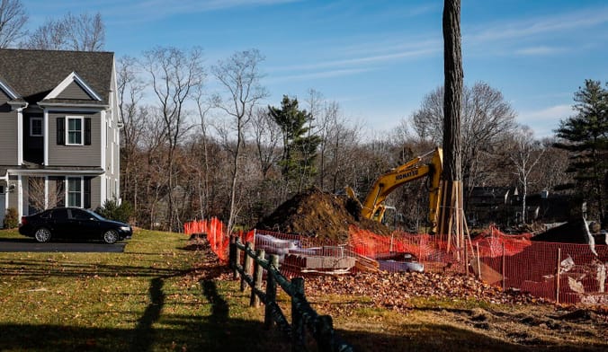 A fence runs between a house with a car parked in front and a patch of land with construction equipment on it