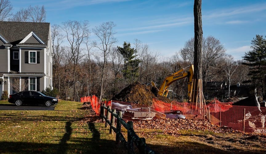 A fence runs between a house with a car parked in front and a patch of land with construction equipment on it