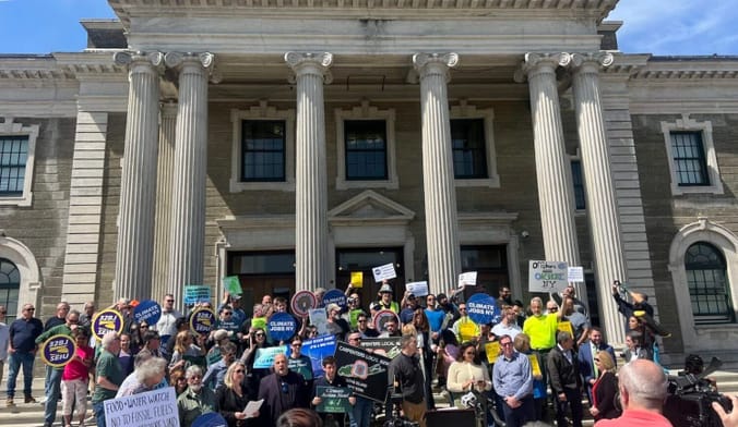 A large group of people stand on the steps of a government building with pillars. Some hold signs supporting offshore wind.